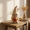Gnomo Ottobre DEKO in ambientazione Japandi Hygge: Su console in legno rustico con candela tealight accesa, spighe di grano e luce autunnale.