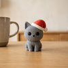 Gomitolo in Grigio, gattino a maglia col cappello di Natale, su un tavolo in legno chiaro